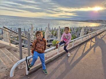 Two children happily pose by a seaside promenade at sunset, with rows of closed umbrellas, calm water, and dramatic clouds creating a peaceful, picturesque atmosphere.