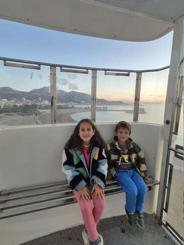 Two smiling children sit inside a Ferris wheel cabin overlooking a scenic beach, with mountains, city buildings, and a beautiful sunset sky in the background.