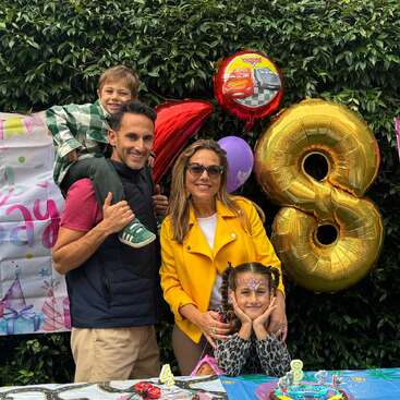 A happy family celebrates a birthday outdoors. Colorful balloons, including a large gold "8", and decorations surround them. The little girl seems to be the birthday child.