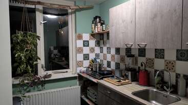 Essa cozinha aconchegante apresenta backsplash de azulejos estampados, plantas penduradas perto de uma janela, prateleiras de madeira com temperos, eletrodomésticos de bancada, utensílios e uma pia de metal brilhante com armários acima.