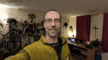 Um homem de óculos e suéter amarelo sorri para uma selfie em uma sala aconchegante, cheia de plantas, com estantes, uma escrivaninha, um laptop e cortinas vermelhas.