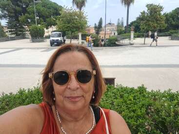 A woman wearing sunglasses and a pearl necklace takes a selfie outdoors. Behind her are green bushes, trees, several people walking, and a parked van.