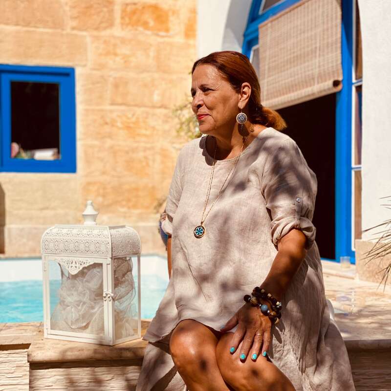 A woman with auburn hair sits by a pool, wearing a beige dress, jewelry, and blue nail polish. Sunlight filters through, creating shadows. Blue-trimmed windows behind her.