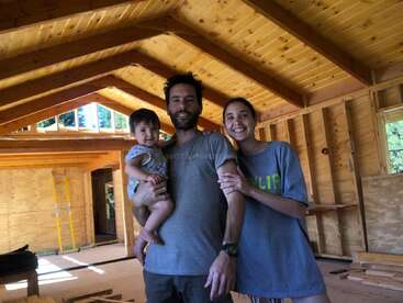 Una sonriente familia de tres miembros se encuentra en el interior de una casa de madera en construcción. Los padres sostienen a su bebé, todos con ropa informal, disfrutando juntos del proyecto de su nuevo hogar.