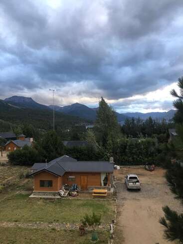 Una pequeña casa de madera se encuentra en una zona rural rodeada de árboles, montañas y nubes espectaculares. Una camioneta está aparcada cerca, creando una escena tranquila y rústica.