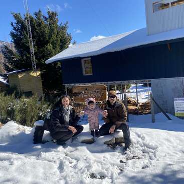 Una familia de tres miembros posa en la nieve delante de una cabaña azul, rodeada de árboles y montañas, disfrutando de un luminoso y soleado día de invierno.