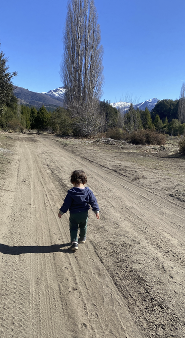 Un niño pequeño camina solo por un camino de tierra rodeado de naturaleza, árboles, montañas y un cielo azul despejado, evocando paz y exploración.