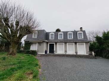 A two-story house with a gray roof, white windows, and a central black door. Leafless trees and greenery surround the building, under a cloudy sky.