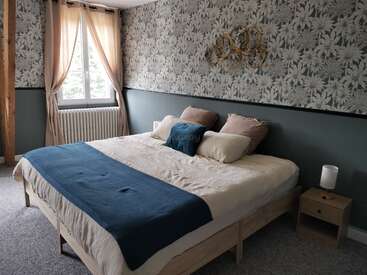 This cozy bedroom features a large bed with neutral bedding and blue accents, floral wallpaper, a wooden nightstand with a lamp, a window, and beige curtains.