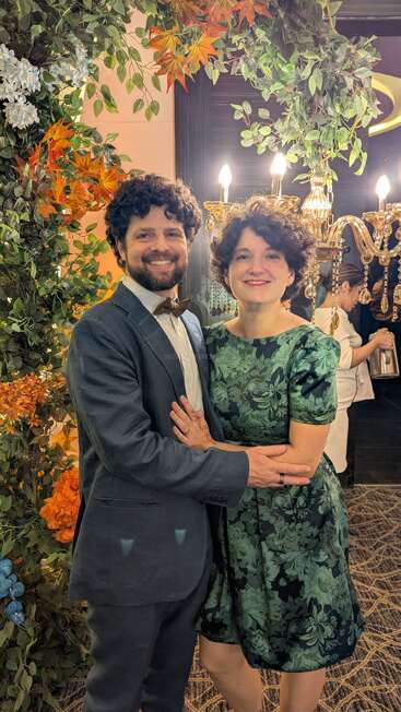 A smiling couple poses at an elegant event, surrounded by greenery and floral decorations, with chandeliers glowing warmly in the background. The atmosphere feels festive and joyful.