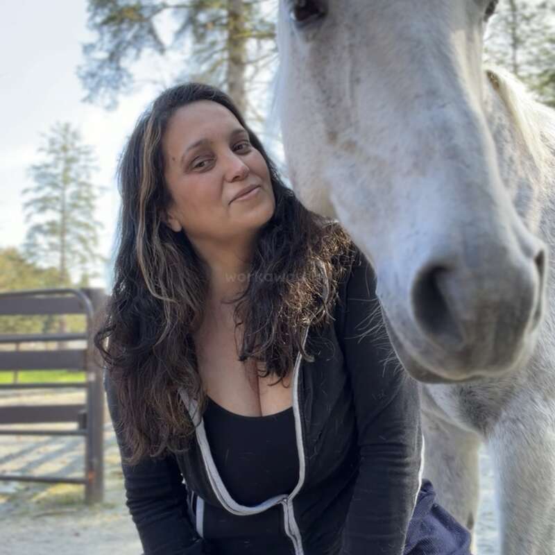 Una mujer de pelo largo y oscuro y camiseta negra posa al aire libre junto a un caballo blanco. Ambos parecen relajados, disfrutando de un día soleado junto a una valla.