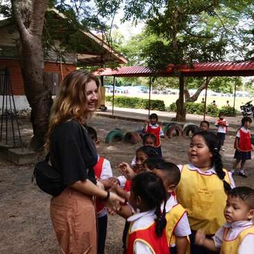 The image depicts a woman in a black shirt and brown pants interacting with a group of children in a playground setting, surrounded by trees and a building.