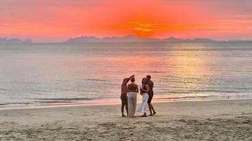 Vier Leute stehen bei Sonnenuntergang an einem Sandstrand und hängen einfach nur zusammen ab. Der leuchtend orangefarbene Himmel spiegelt sich im ruhigen Meer und sorgt für eine entspannte Atmosphäre.