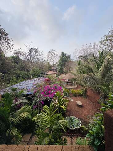 A lush tropical garden filled with vibrant flowers and palm trees surrounds rustic thatched huts. A small pond adds tranquility under a partly cloudy blue sky.