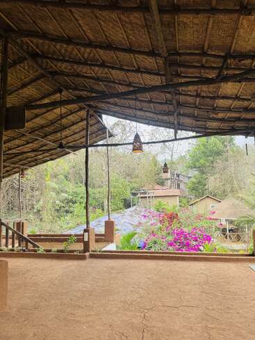 A rustic, open-air structure with a thatched roof overlooks a lush garden filled with colorful flowers, greenery, and distant houses nestled among trees. Peaceful atmosphere.