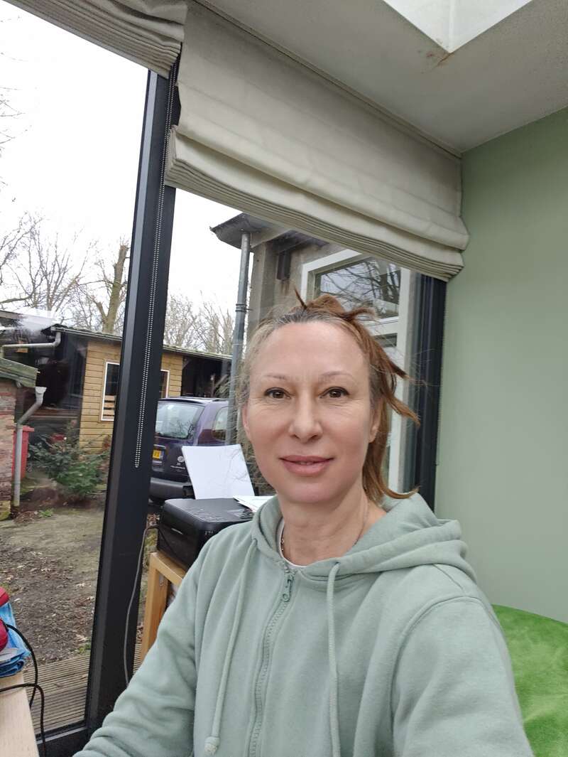 A woman in a light green hoodie smiles indoors by a large window with a garden view. Papers, a printer, and outdoor buildings are visible behind her.