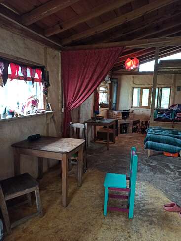 Ein gemütlicher, rustikaler Raum mit Holzmöbeln, bunten Vorhängen, einem grünen Stuhl und freigelegten Holzdecken. Die warme Beleuchtung und die heimelige Atmosphäre lassen auf einen lebendigen, kreativen Raum schließen.