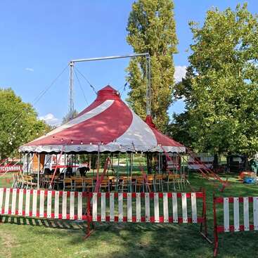 Uma tenda de circo vermelha e branca fica em um gramado cercado por árvores e barreiras listradas. Cadeiras vazias estão dispostas dentro dela, esperando os visitantes sob um céu azul claro.