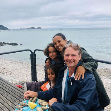 La imagen muestra a una familia de cuatro miembros sentados a una mesa de madera con un plato de ostras, con el telón de fondo de una playa serena y el océano.