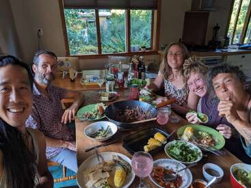 Cinq amis sont réunis autour d'une table en bois, partageant un repas coloré fait maison, souriant et appréciant la compagnie des autres dans une cuisine confortable et ensoleillée avec vue sur le jardin.