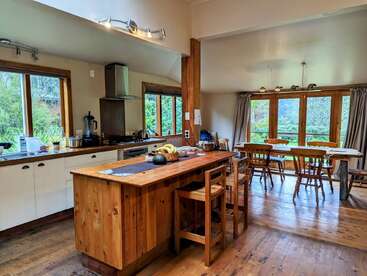 Cette cuisine et cette salle à manger sont dotées de sols en bois, d'un îlot central, d'armoires blanches, de grandes fenêtres, d'une lumière naturelle, de verdure à l'extérieur et d'un mobilier en bois confortable.