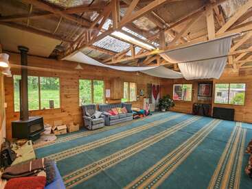 Cette pièce spacieuse et rustique se caractérise par des murs en bois, des poutres apparentes, un poêle à bois, des canapés confortables, une moquette verte, de grandes fenêtres, des rideaux en tissu au plafond et des éléments décoratifs partout.