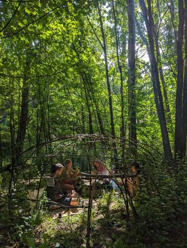 Quatre personnes sont assises ensemble à l'intérieur d'un cadre en bois en forme de dôme dans une forêt luxuriante et ensoleillée, entourées de verdure, partageant un moment paisible et intime en plein air.
