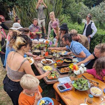 Un groupe de personnes se réunit autour d'une table en bois en plein air, partageant un festin coloré composé de plats variés, de salades fraîches, de fruits et de fleurs, et profitant d'une conversation animée.