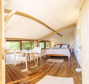 Dieses gemütliche, sonnendurchflutete Schlafzimmer verfügt über eine Holzdecke mit sichtbaren Balken, ein bequemes Doppelbett, einen kleinen Esstisch mit Stühlen, große Fenster und einen warmen Holzboden.
