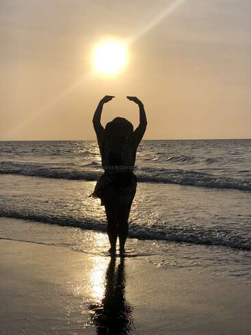 Una persona está de pie en la playa al atardecer, silueteada contra el sol resplandeciente, con los brazos levantados para enmarcar la luz, con suaves olas y reflejos debajo.