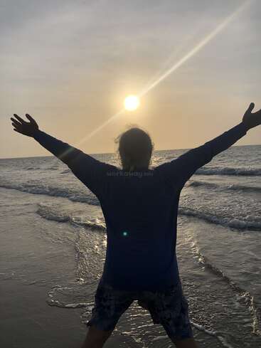 Una persona está de pie en la playa con los brazos extendidos, frente a la puesta de sol sobre el mar, abrazando la serena belleza y la libertad del impresionante momento de la naturaleza.