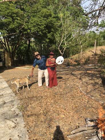 Un hombre y una mujer, ambos con sombrero, están de pie fuera con un perro. La mujer sostiene un tambor blanco. Se ven árboles y una pequeña hoguera.