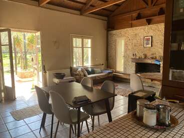 Ce salon-salle à manger chaleureux et baigné de soleil est doté d'un plafond en bois, d'une cheminée en pierre, d'un mobilier moderne et de grandes fenêtres. La porte ouverte dévoile une vue sur un extérieur verdoyant et luxuriant.