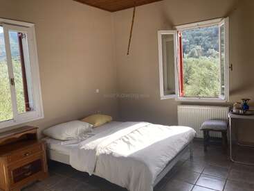 Cette chambre lumineuse et sobre est dotée d'un lit bien fait, d'une table de chevet en bois, d'un sol carrelé, de deux fenêtres ouvertes offrant une vue panoramique sur les montagnes, ainsi que d'une décoration minimaliste qui crée une atmosphère paisible.