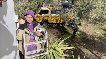 Un homme vêtu d'un sweat à capuche et d'une veste violets est debout sur une échelle, en train de travailler près d'un mur. Derrière lui, une camionnette jaune est garée au milieu des oliviers.