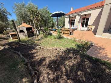 Une terrasse chaleureuse, agrémentée de meubles en palettes et d'un grand parasol, jouxte une maison aux tuiles en terre cuite, entourée d'arbres, de plantes et d'un four à briques extérieur.