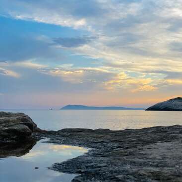 The image depicts a serene rocky shoreline with a tranquil body of water, set against a vibrant sky with clouds and a distant island or mountain range.
