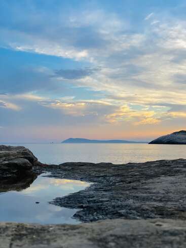 The image depicts a serene rocky shoreline with a tranquil body of water, set against a vibrant sky with clouds and a distant island or mountain range.