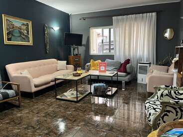 L'image représente un salon aux murs sombres et au sol carrelé marron, avec un canapé beige, une table basse et une fenêtre aux rideaux blancs.