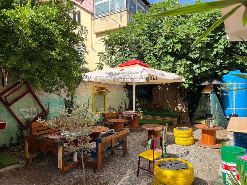 L'image représente un espace extérieur serein avec des tables et des chaises en bois, entouré d'une végétation luxuriante et d'un parasol blanc, dégageant une atmosphère chaleureuse et accueillante.