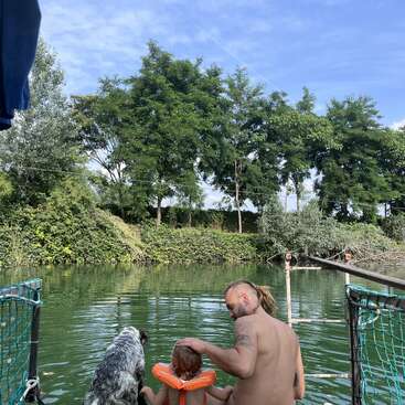 A man, a child with floaties, and a dog sit on a dock by a green river, surrounded by lush trees and a clear blue sky.
