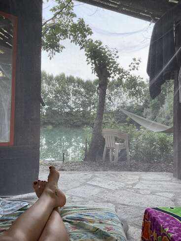 A person relaxes indoors with crossed legs on a bed, gazing out at a tranquil lakeside view, trees, hammock, and chair under soft daylight.