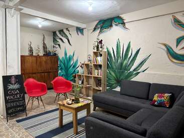 Ce salon confortable de l'auberge de jeunesse est aménagé avec un canapé gris foncé, deux fauteuils rouges, des fresques représentant des agaves, une bibliothèque, une décoration colorée, des meubles en bois et un panneau d'accueil écrit à la craie.