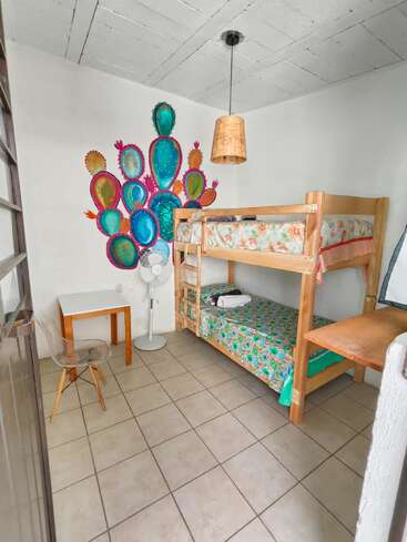 Cette chambre chaleureuse est équipée d'un lit superposé en bois avec une literie à motifs floraux, d'un petit bureau, d'une chaise transparente, d'un ventilateur sur pied et d'une fresque murale colorée représentant des cactus.