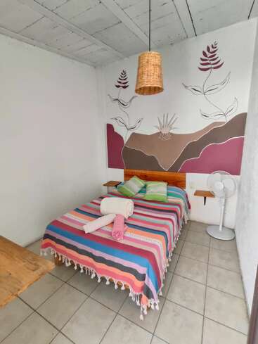Cette chambre chaleureuse est agrémentée d'un couvre-lit à rayures colorées, de deux coussins verts, d'une décoration murale représentant des feuilles, d'une lampe en osier, d'un ventilateur, d'un sol carrelé et d'un mobilier minimaliste.