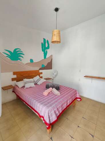 Voici une chambre confortable équipée d'un lit double, d'un couvre-lit rouge, d'oreillers, d'une fresque murale sur le thème du désert, d'un plafonnier, d'un ventilateur et d'une décoration minimaliste sur un sol carrelé.