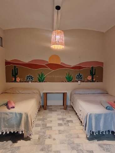 Cette chambre chaleureuse comprend deux lits recouverts de couvertures colorées, une table centrale en bois, un éclairage tamisé et une fresque murale représentant un coucher de soleil sur des montagnes désertiques et des cactus.