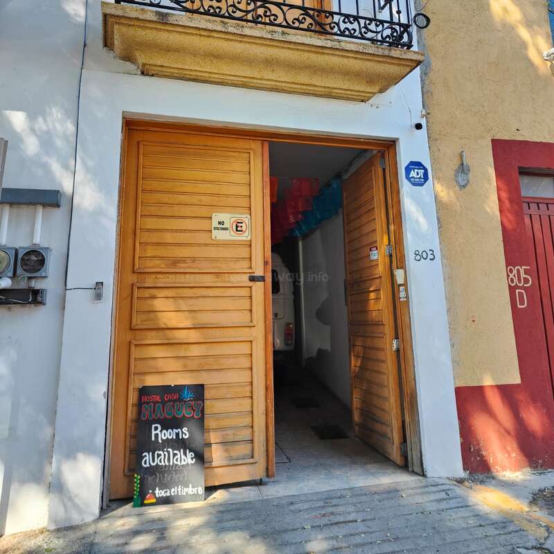 Une porte en bois donnant sur une auberge dont l'entrée est ouverte, un panneau en ardoise indiquant les chambres disponibles et des décorations colorées à l'intérieur. Le numéro 803 est inscrit sur le mur du bâtiment.