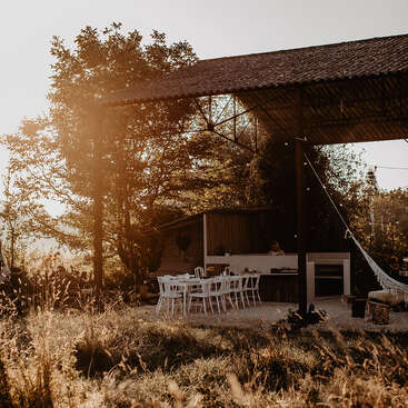 Golden sunlight bathes an outdoor dining area with white chairs, under a rustic shelter. Trees, tall grass, and a hammock create a peaceful countryside scene.