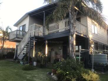The image depicts a two-story house with a balcony, stairs, and a water tank, surrounded by lush greenery and palm trees in a serene outdoor setting.